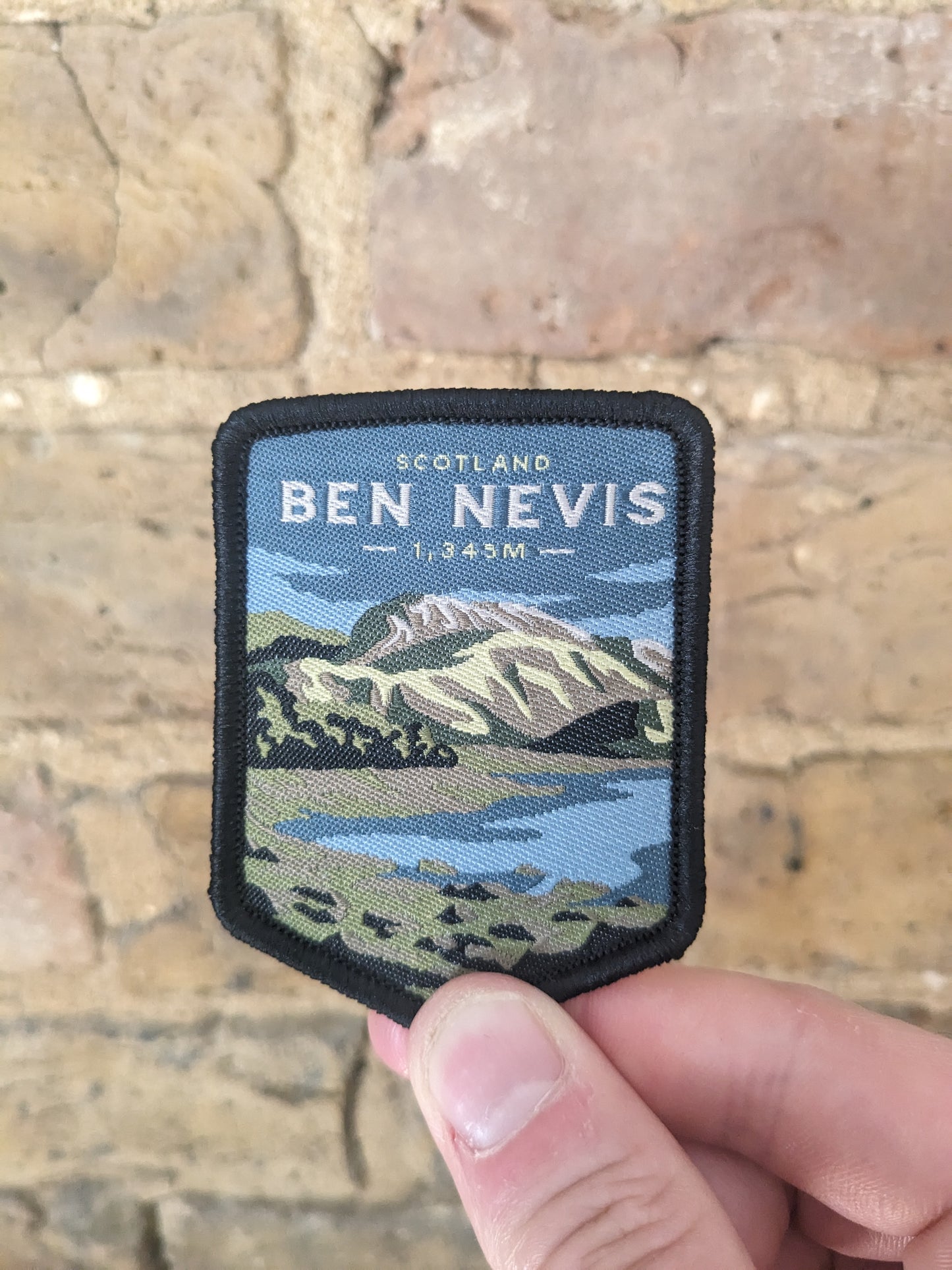 Ben Nevis summit patch held up against a brick background. The badge features the iconic mountain with a blue background.