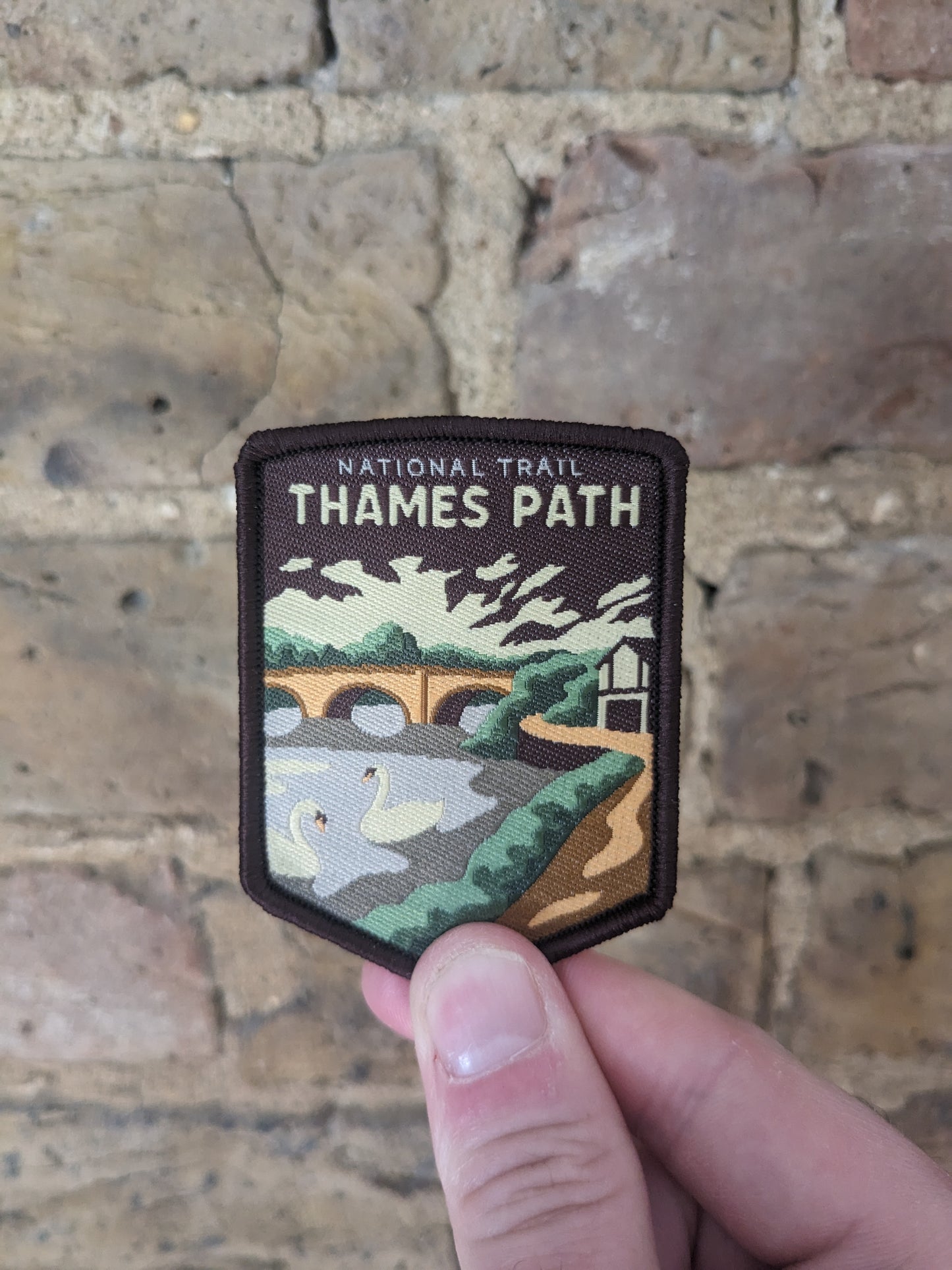 image of the badge for the Thames Path featuring a swan on the river against a backdrop of a train bridge (a common sight on the thames path), the badge being held up against a wall backdrop