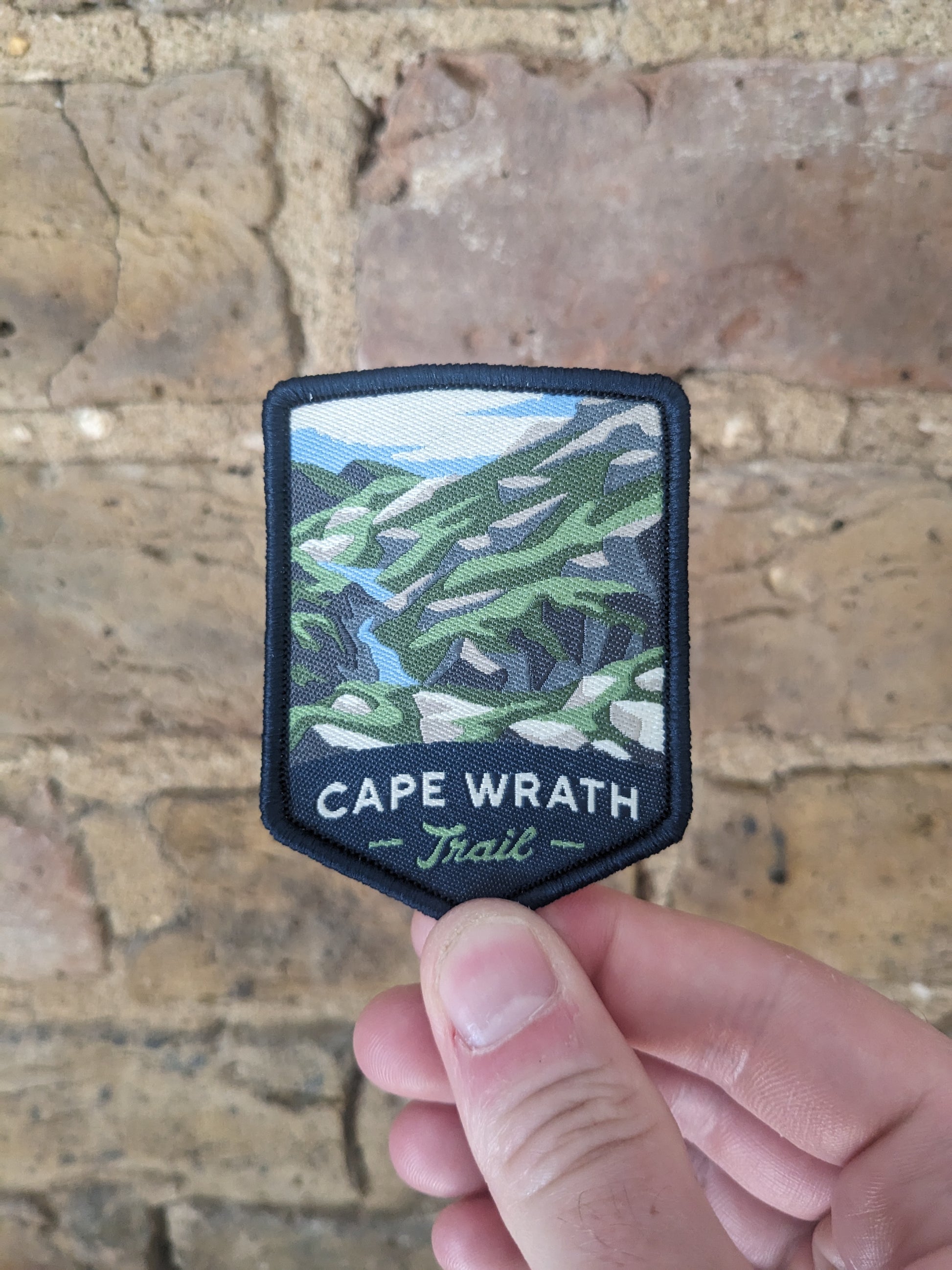 Image of the Cape Wrath Trail badge being held against a backdrop of a brick wall.