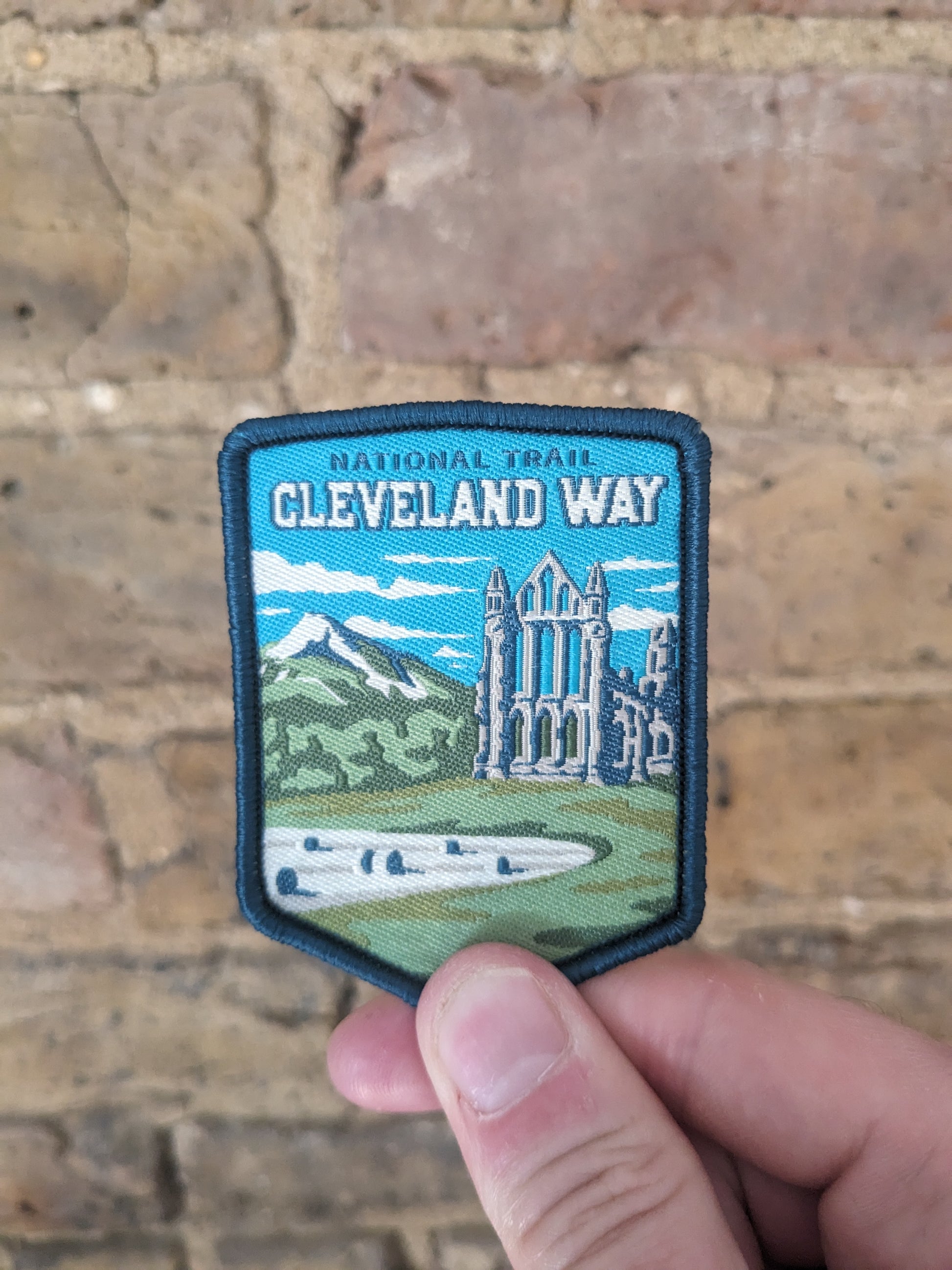 The Cleveland Way patch/badge being held up against a brick wall background. The badge has a blue background and features Roseberry Topping and the ruins of an Abbey on the trail.