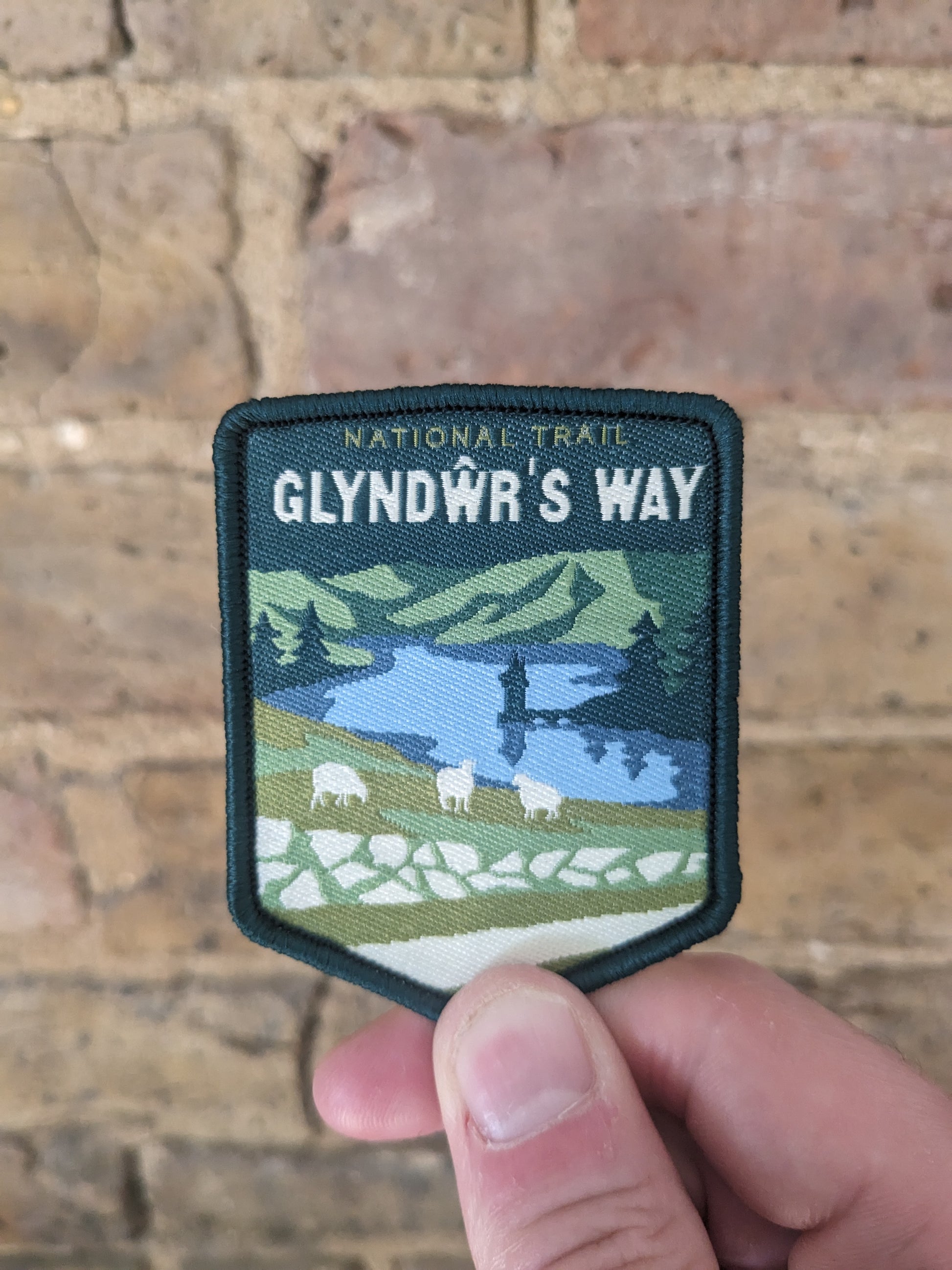 Picture of the Glyndwrs Way patch being held against a brick wall backdrop. The patch features a view towards Lake Vyrnwy