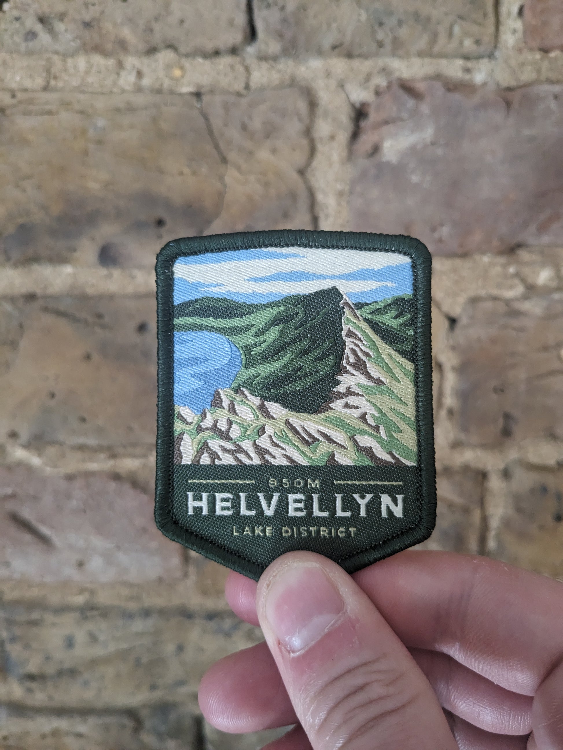 Image of a Helvellyn badge depicting the Striding Edge route, being held against a brick wall backdrop.