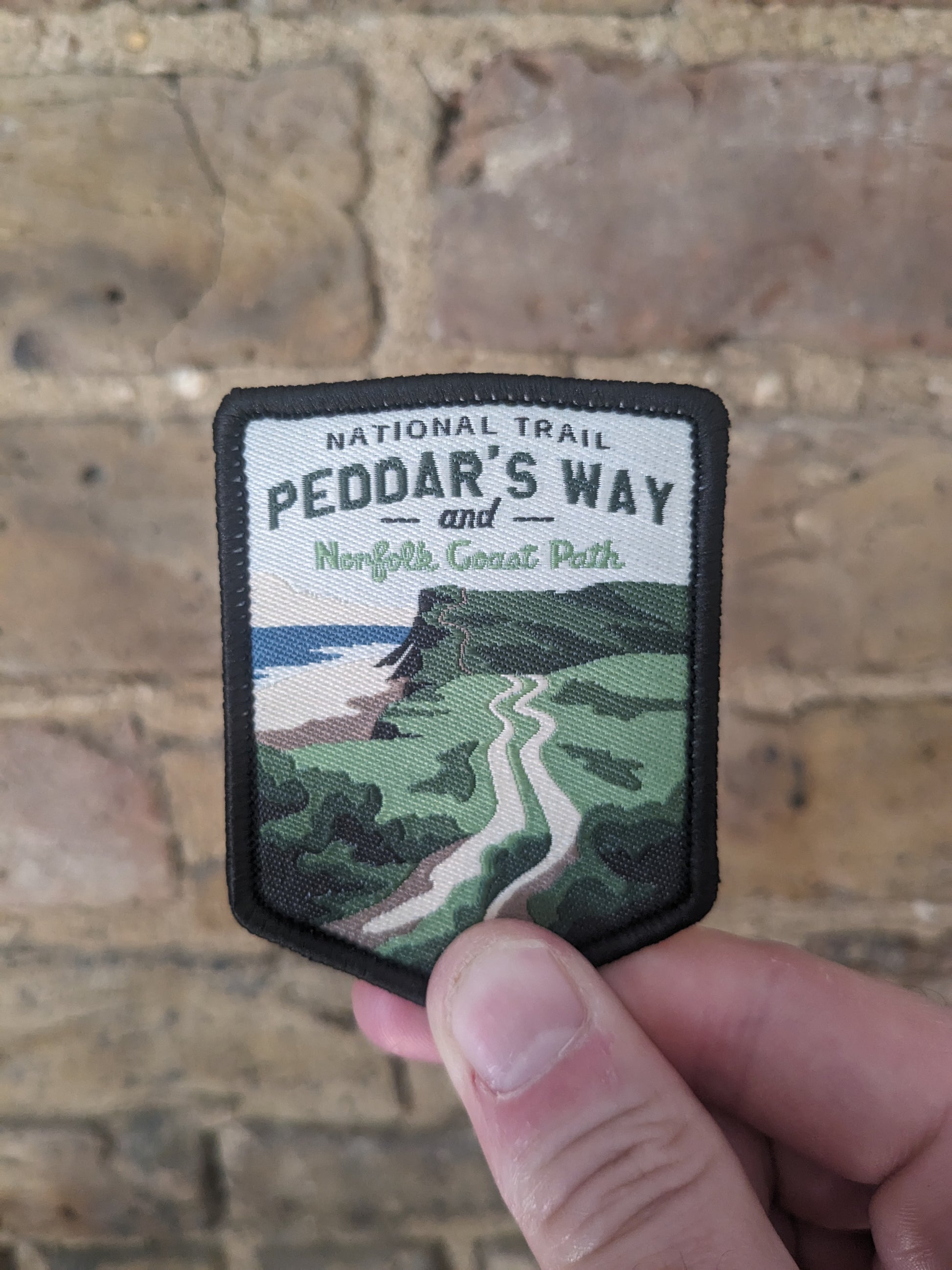 Picture of the Peddars Way and Norfolk Coast Path patch being held against a brick backdrop. The patch features a classic coastline view from the trail.