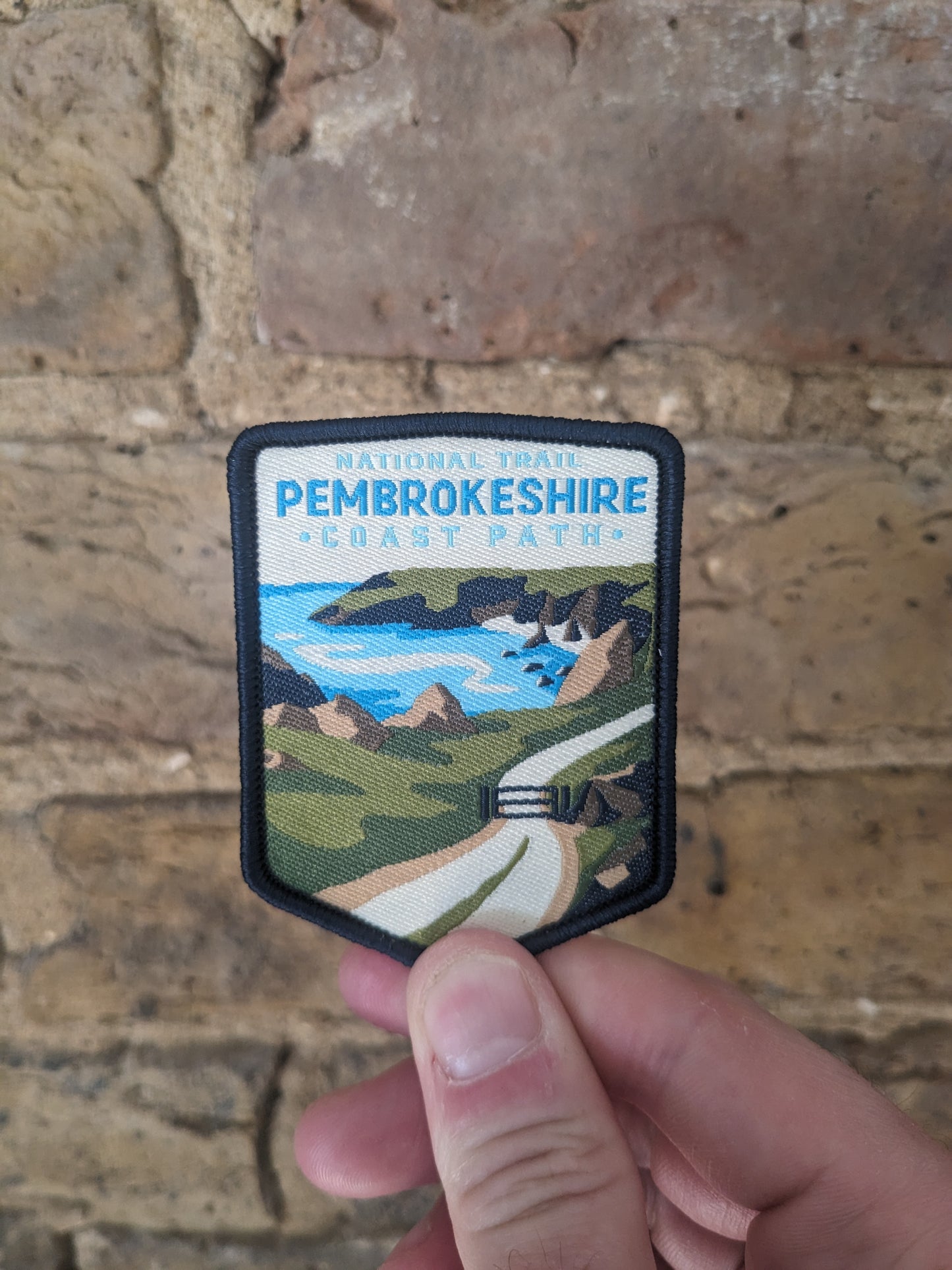 Picture of the Pembrokeshire Coast Path National Trail patch depicting the Welsh coastline being held up against a brick wall