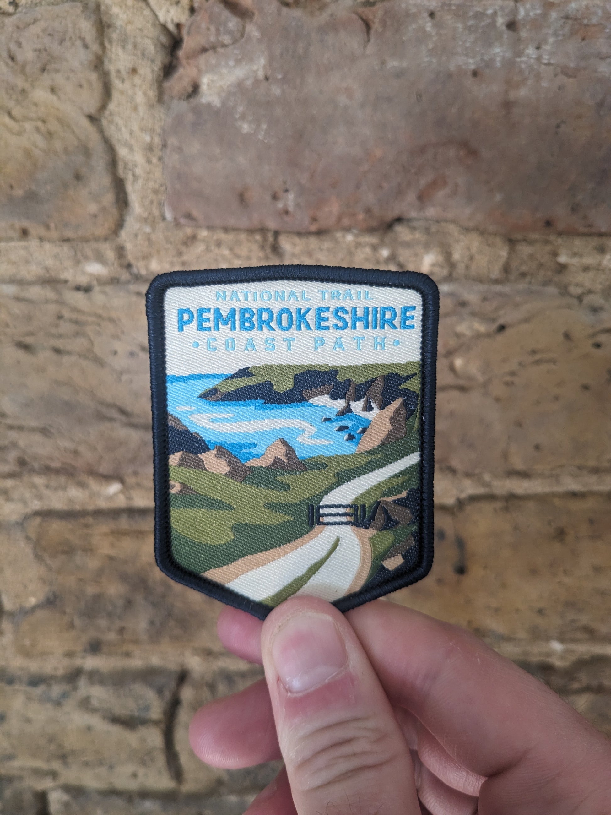 Picture of the Pembrokeshire Coast Path National Trail patch depicting the Welsh coastline being held up against a brick wall