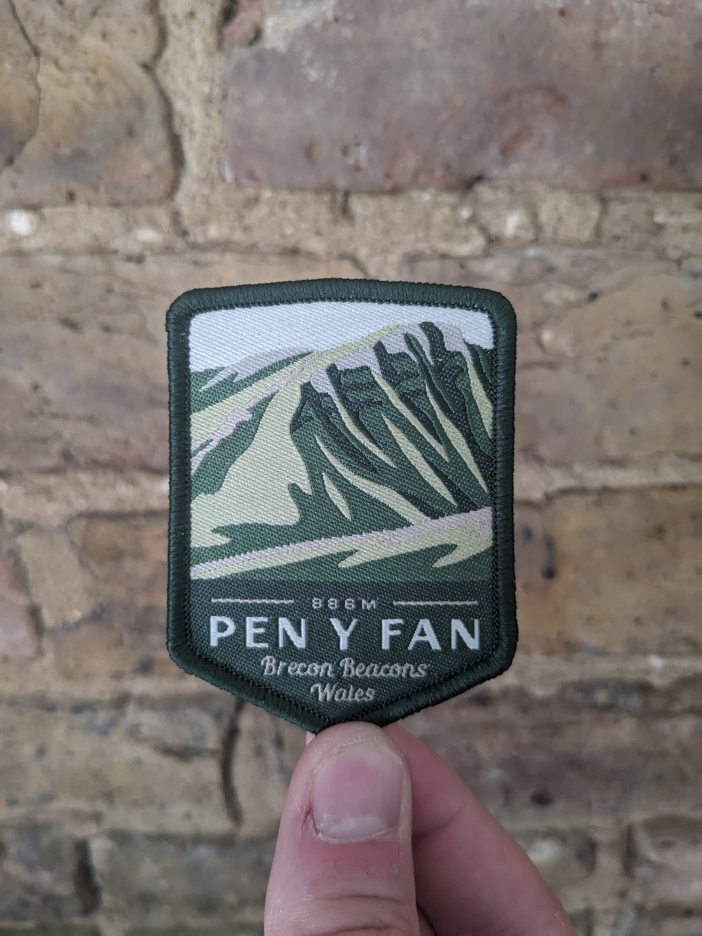 Pen y Fan Badge held against the backdrop of a brick wall. The patch itself features the iconic mountain in the distance.