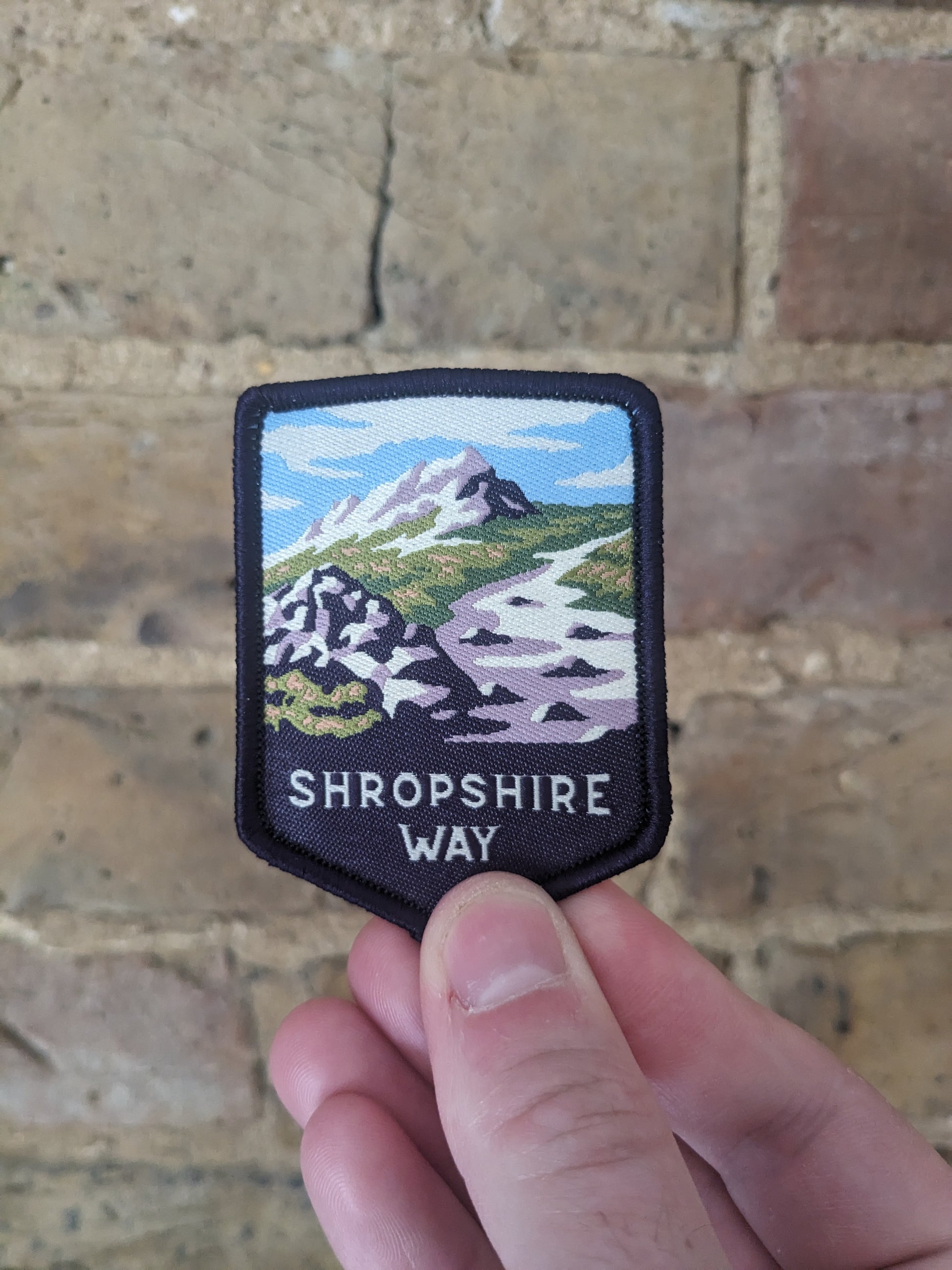 Image depicting the Shropshire Way patch held up against a brick wall. The patch itself features the Stiperstones hill.