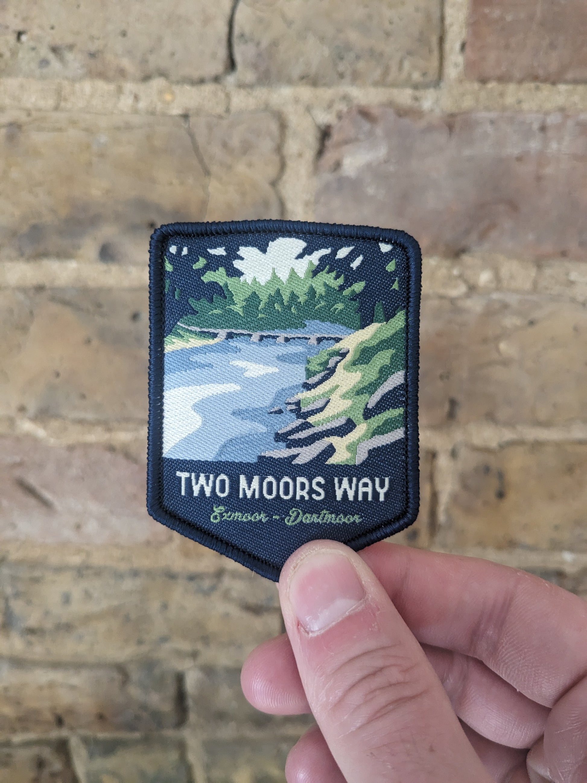 Image of the Two Moors Way patch being held up against a brick wall. The patch depicts the Tarr Steps on the River Barle